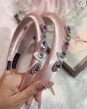 A hand holds two pink satin headbands decorated with pink and silver rhinestones and small white bows, against a soft pink background with a gift box and lace accessory visible.
